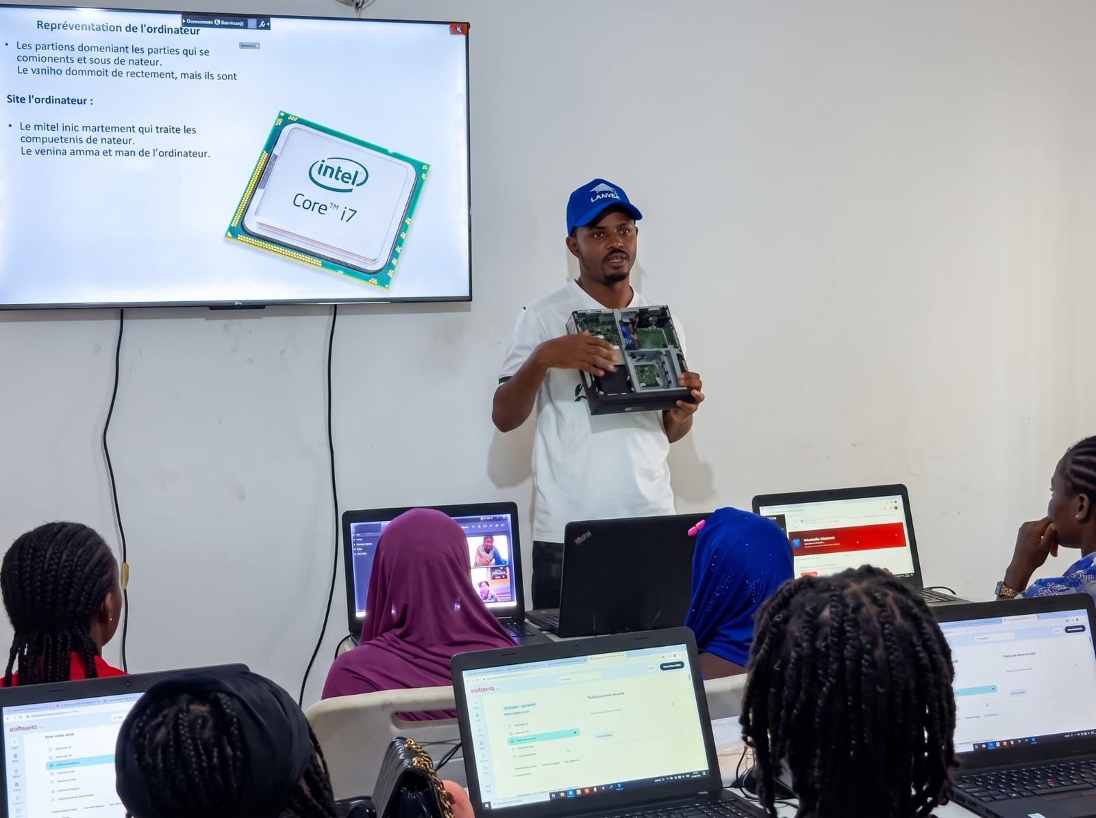 A facilitator demonstrates computer hardware to young learners using laptops in a community classroom.